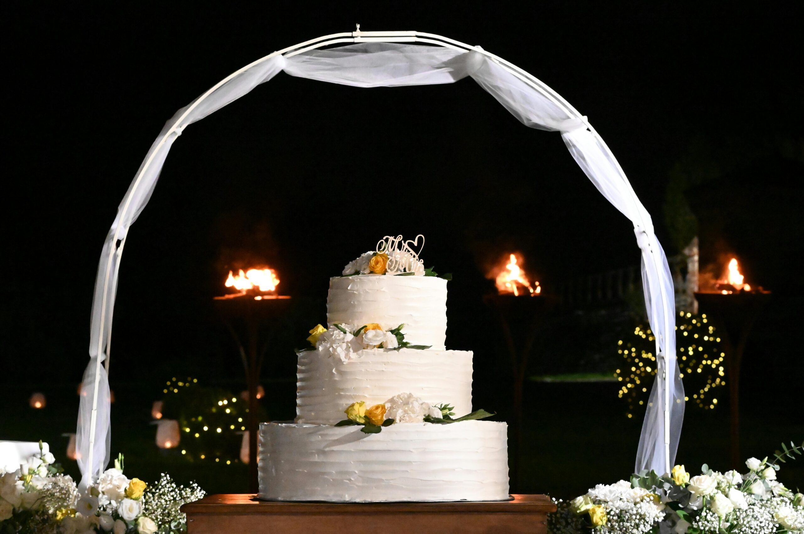 Beautiful white wedding cake under arch at night with fire torches in background.